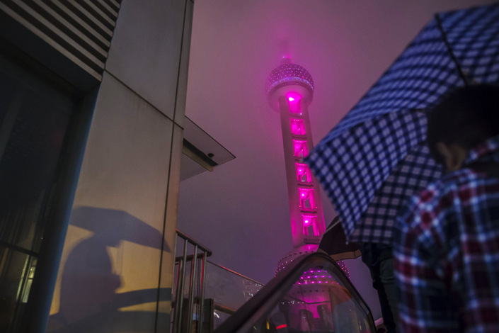 A pedestrian holding an umbrella rides up an escalator as the Oriental Pearl Tower stands illuminated at night in the Lujiazui district of Shanghai