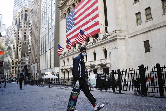 A boy wearing a mask and carrying a skateboard walks past an American flag on Wall Street on March 24. Photo: People.vcg