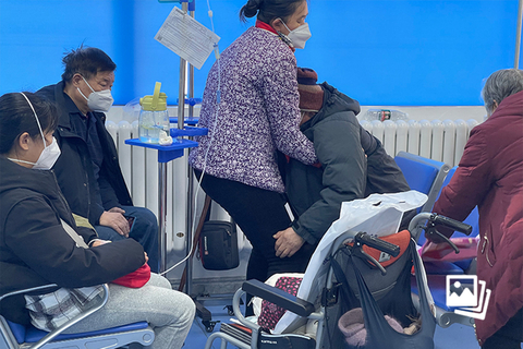 2022年12月28日，北京306医院发热门诊输液室内，一位陪护亲属将病人转移到轮椅上，推着他到有吸氧、雾化治疗设备的输液座位上去。近期，北京市仍面对疫情冲击，多家医院发热门诊及急诊病患数量较多，病患床位仍紧缺，医护人员加班成常态。图：海杜马（特约）